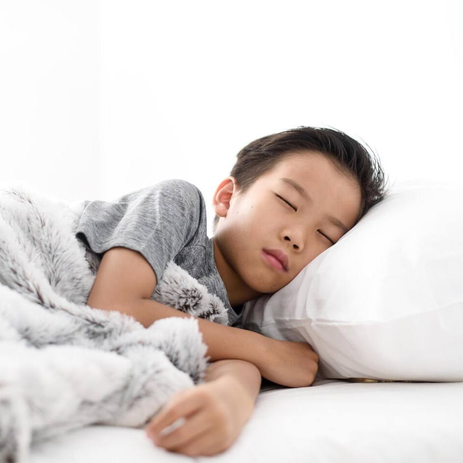 一个穿着灰色睡衣的小男孩的肖像，睡在白色的床单和枕头上，下面是柔软的模糊灰色毯子。