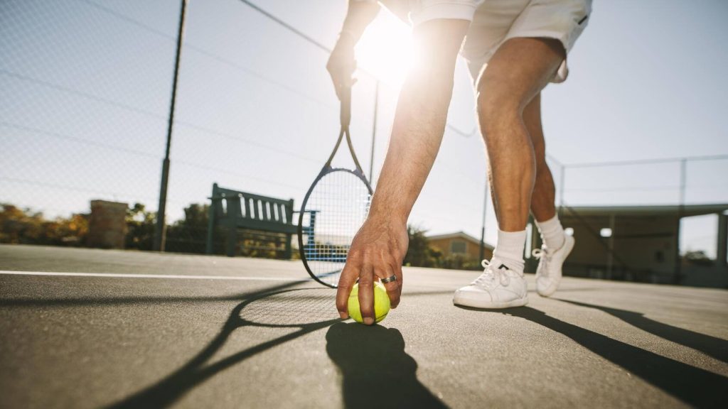 Vista de ángulo bajo de un jugador de tenis que se inclina hacia adelante para recoger la pelota con el sol en el fondo.