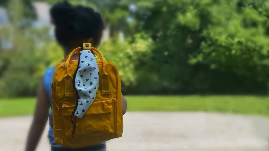 Una vista trasera de una niña esperando afuera, lista para ir a la escuela. Lleva su mochila amarilla a la que está asegurada su mascarilla protectora.